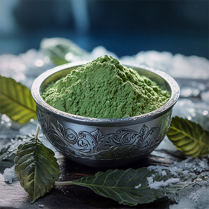 A close-up of green kratom powder in an ornate bowl surrounded by kratom leaves on a wooden surface